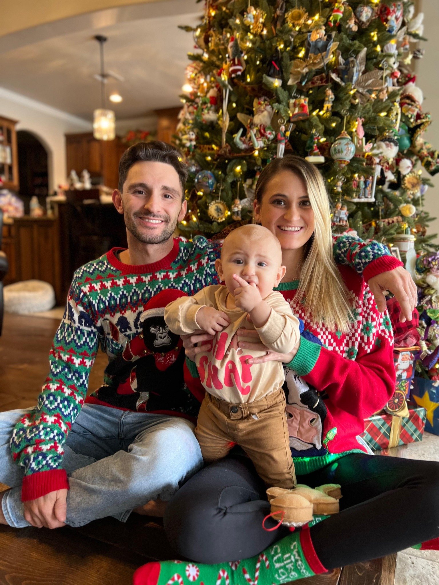 Christmas photo of husband, wife, and a baby in front of a Christmas tree.